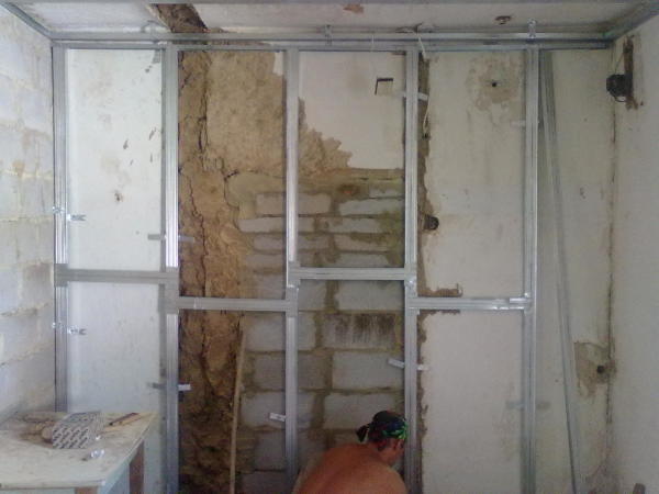 The photo shows the process of mounting a metal frame for wall sheathing with plasterboard sheets to align it